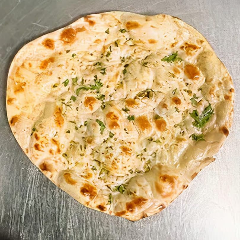 Golden-brown Indian naan bread, speckled with green herbs, rests on a metallic surface.